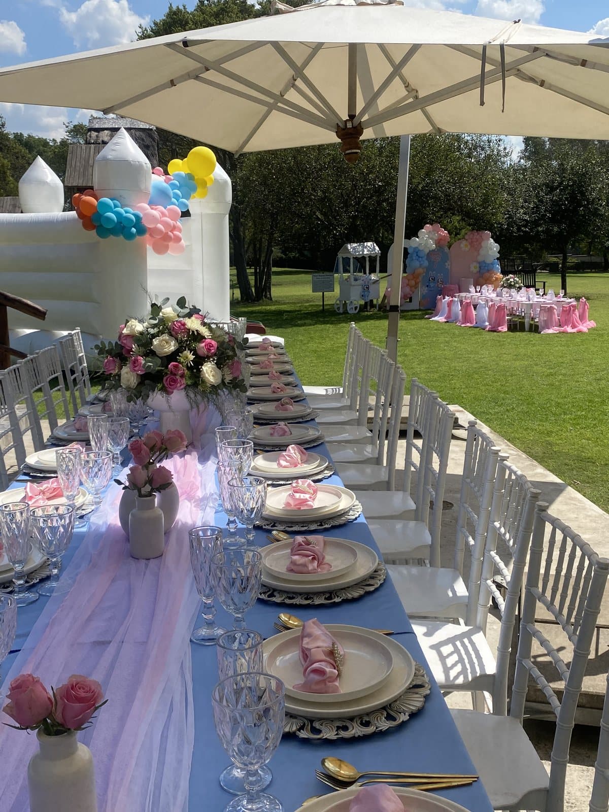 Outdoor long table with blue tablecloth and pastel balloon arch under an umbrella