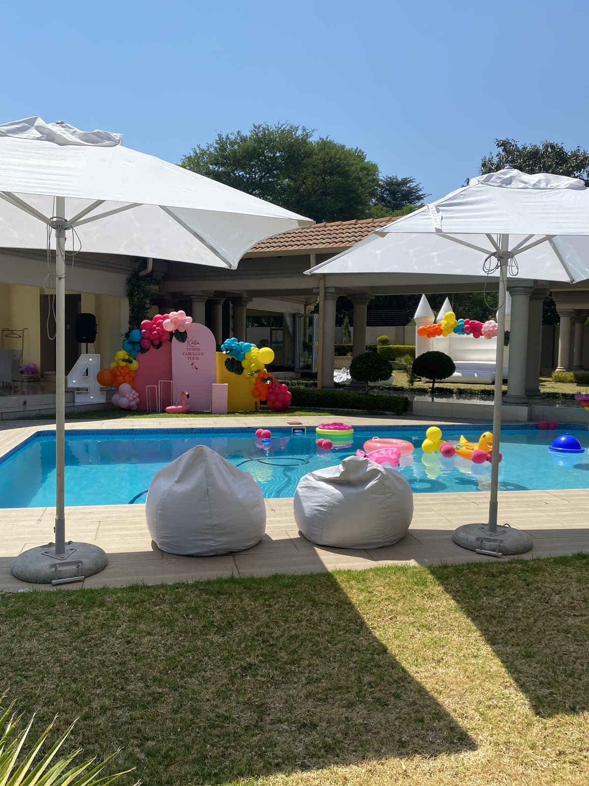 Poolside party setup with white bean bags, umbrellas and scattered pastel balloons