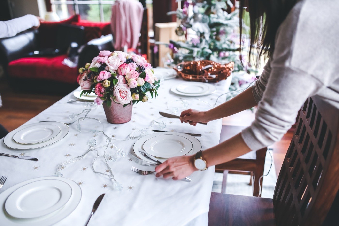A woman sets a dinner table at home with white and gold place settings and pink flowers